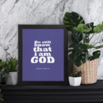 Be Still and Know That I Am God Psalm 46:10 Christian Wall Art with bold white text on a dark purple background. Wall Art displayed on the table in a black frame