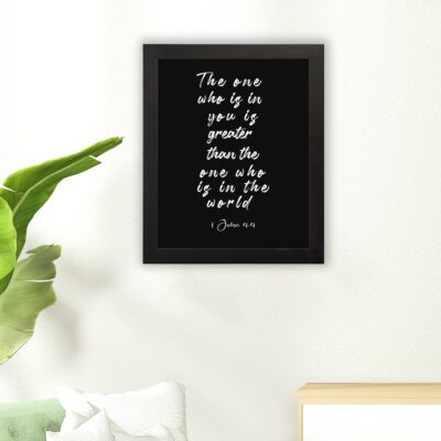 Black wall art with white text displaying The one who is in you is greater than the one who is in the world 1 John 4:4. hung on the floor in a black frame