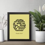 Infidu The Lord Of Hosts Is With Us Psalm 46:7 Bible Verse Wall Art on light yellow background. displayed on the table in a black frame