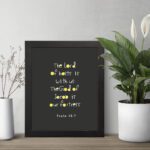 Psalm 46:7 Bible Verse Christian Wall Art - The Lord of Hosts is With Us, Minimalistic Design with Gold Accents. displayed on the table in a black frame