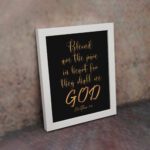 Infidu wall art with the text Blessed is the pure in heart, for they shall see God in gold font on a black background. displayed on the wall in a white frame