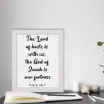 The Lord Of Hosts Is With Us - Psalm 46:7 Bible Verse Wall Art with White Text on Light Background Framed in Black Wall Art displayed on the table in a white frame