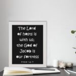 The Lord Of Hosts Is With Us - Psalm 46:7 Bible Verse Wall Art with White Text on Black Background Wall Art displayed on the table in a white frame