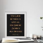 The Lord Of Hosts Is With Us - Psalm 46:7 Bible Verse Wall Art with Golden Text on Black Background Wall Art displayed on the table in a white frame