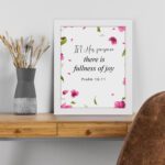 Psalm 16:11 wall art with a white background and pink flower petals, featuring the text 'In His presence there is fullness of joy. Wall Art displayed on the wall in a white frame