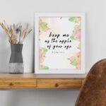 Keep Me As The Apple Of Your Eye Psalm 17:8 Bible Verse Wall Art with apple and leaf design, soft pastel colors, and white background. Wall Art displayed on the table in white frame