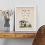 Psalm 17:8 wall art with a soft cursive font and a peaceful illustration of a person sitting under a tree. Wall Art is displayed on the table in a white frame.