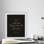 Black wall art with gold text saying SHE IS FAR MORE PRECIOUS THAN JEWELS from Proverbs 31:10, styled with decorative elements. displayed on the table in a white frame