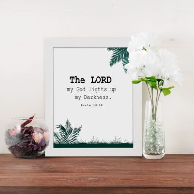 The Lord My God Lights Up My Darkness Psalm 18:28 Bible Verse Wall Art with black text and green tropical leaves on a white background. Wall Art displayed on the table in a white frame