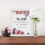 The Lord My God Lights Up My Darkness Psalm 18:28 Bible Verse Wall Art with black text and bold red, orange, blue, and yellow brush strokes on a white background. Wall Art displayed on the table in a white frame