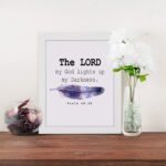 The Lord My God Lights Up My Darkness Psalm 18:28 Bible Verse Wall Art with black text and a purple and blue feather on a white background. Wall Art displayed on the table in a white frame