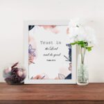 Trust In The Lord And Do Good Psalm 37:3 Bible Verse Wall Art with watercolor and floral designs on a white background. Wall Art displayed on the table in a white frame