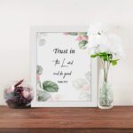 Trust in the Lord and Do Good Psalm 37:3 Bible Wall Art with botanical green leaves and pink flowers on a white background. Wall Art displayed on the table in a white frame