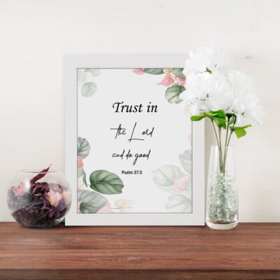 Trust in the Lord and Do Good Psalm 37:3 Bible Wall Art with botanical green leaves and pink flowers on a white background. Wall Art displayed on the table in a white frame