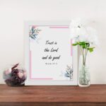 Infidu Bible wall art featuring Psalm 37:3 in black text with blue and pink floral accents and a soft pink border. Wall Art displayed on the table in a white frame