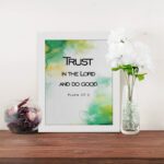 Trust in the Lord and Do Good Psalm 37:3 Bible Verse Wall Art with watercolor green and yellow background. Wall Art displayed on the table in a white frame