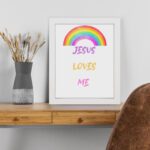 Infidu Jesus Loves Me Bible Wall Art with rainbow arc and colorful text in purple, yellow, and pink fonts Wall Art displayed on the table in a black frame