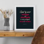 Infidu Psalm 46:1 Bible Wall Art with a deep blue background, pink and white text, featuring God is our refuge and strength. Wall Art displayed on the table in a white frame