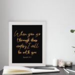 Black wall art with gold cursive text reading When you go through deep waters I will be with you from Isaiah 43:2. displayed on the table in a white frame