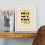 Psalm 50:15 Bible Wall Art with bold black, orange, and yellow text and simple floral embellishments on a cream background. Wall Art displayed on the table in a white frame