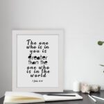 White wall art with black text reading The one who is in you is greater than the one who is in the world 1 John 4:4. displayed on the table in a white frame