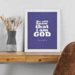 Be Still and Know That I Am God Psalm 46:10 Christian Wall Art with bold white text on a dark purple background. Wall Art displayed on the table in a white frame