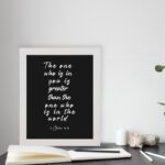 Black wall art with white text displaying The one who is in you is greater than the one who is in the world 1 John 4:4. displayed on the table in a white frame