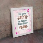 Christian wall art with brown 'Faith' and 'Fear' text, pink leaves, and a light pink floral background. Wall Art displayed on the wall in a white frame