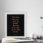 1 John 4:4 Bible verse wall art in gold text on a black background, featuring the quote "The one who is in you is greater than the one who is in the world. displayed on the table in a white frame