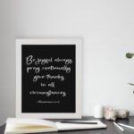 Bible verse wall art featuring 1 Thessalonians 5:16-18 in white cursive text on a solid black background, arranged clearly for easy reading. displayed on the table in a white frame