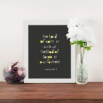 Psalm 46:7 Bible Verse Christian Wall Art - The Lord of Hosts is With Us, Minimalistic Design with Gold Accents. displayed on the table in a white frame