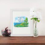 Psalm 46:7 Bible verse wall art with serene watercolor landscape of trees, hills, and water, featuring the verse in black text. displayed on the table in a white frame