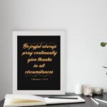 Bible verse wall art featuring 1 Thessalonians 5:16-18 in gold cursive text on a solid black background, arranged in a stacked format. displayed on the table in a white frame