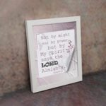 Zechariah 4:6 Bible verse wall art with soft purple text, bold LORD, decorative plant motif, and light purple folded paper background. displayed on the wall in a white frame