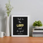 Psalm 37:3 Bible verse wall art with a black background, Lord highlighted in yellow. Christian home decor by Infidu. Wall Art displayed on the table in a black frame