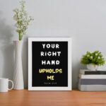 Infidu Your Right Hand Upholds Me Psalm 63:8 Bible Wall Art with a black background, yellow, and white font, and white frame displayed on the table