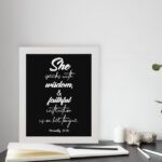 Proverbs 31:26 Bible wall art with white text on a plain black background, featuring cursive and regular fonts emphasizing "She" and "wisdom." displayed on the table in a white frame