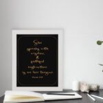 Proverbs 31:26 Bible wall art with gold text on a black background, framed by a golden border. Featuring elegant cursive font. displayed on the table in a white frame