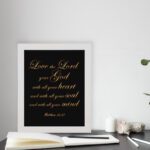 Matthew 22:37 Bible wall art with gold text on a black background, framed in black, reading Love the Lord your God. displayed on the table in a white frame