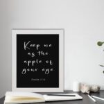 Keep Me As The Apple Of Your Eye Psalm 17:8 white cursive text on black background - Christian wall art for home decor displayed on the table in a white frame