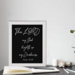 The Lord My God Lights Up My Darkness Psalm 18:28 white handwritten text on black background Christian wall art displayed on the table in a white frame