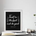 Trust In The Lord And Do Good Psalm 37:3 white text with decorative branches on black background Christian wall art displayed on the table in white frame