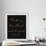 Trust In The Lord And Do Good Psalm 37:3 golden text on black background Christian wall art displayed on the table in a white frame