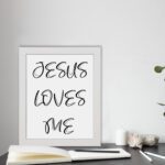 Jesus Loves Me Bible wall art with black script text on a white background. Wall Art displayed on the table in a white frame