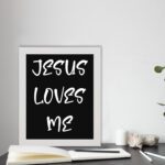 Jesus Loves Me Bible wall art with bold gold text and a decorative gold border on a black background. Wall Art displayed on the table in a white frame