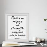 Psalm 46:1 Bible wall art with black bold text on a white background, saying 'God is our refuge and strength, a very present help in trouble. Wall Art displayed on the table in a white frame