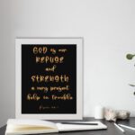 Psalm 46:1 Bible wall art with gold handwritten text on a black background, saying 'God is our refuge and strength. Wall Art displayed on the table in a white frame