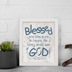 Infidu Matthew 5:8 Christian wall art with blue text on a white background, featuring a subtle pastel floral design and stylized GOD. Wall Art displayed on the table in a white frame