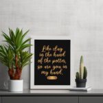 Infidu wall art with the text Like clay in the hand of the potter, so are you in my hand in golden letters on a black background. displayed on the table in a white frame