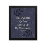 Purple-blue background with black leaf design, white text 'The Lord My God Lights Up My Darkness Psalm 18:28 Bible Verse Wall Art black frame hung on the wall.